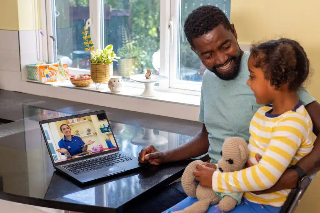 A man and a little girl sitting at a table with a laptop and a teddy bear.