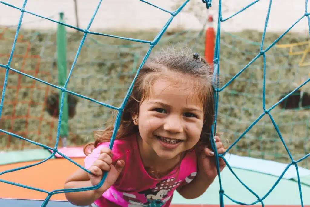 A little girl smiling through a net in a playground.